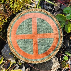 Op de afbeelding: Een ronde, groene en oranje stoffen patch met een geweven ontwerp. De patch heeft een bruine rand en een geweven kruis in het midden.