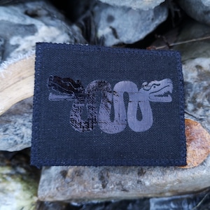 Könnte beinhalten: Schwarzer Stoff-Patch mit einem schwarzen Schlangen-Design. Die Schlange ist zusammengerollt und hat einen Kopf mit einem scharfen Maul.