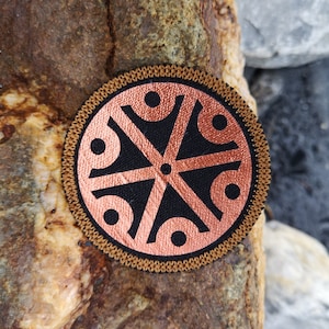 May include: A round, brown patch with a black and copper design. The design features a star-like shape with eight points, each point having a circle at the end. The patch is surrounded by a brown stitched border.