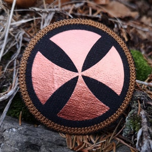 Op de afbeelding: Een ronde, bruine, geborduurde patch met een koperkleurig kruisontwerp. Het kruis heeft vier punten en is gecentreerd op een zwarte achtergrond.