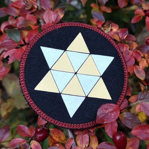 Puede incluir: Un parche bordado negro y rojo con un diseño de estrella geométrica amarilla y blanca.