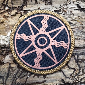 May include: A round, black fabric patch with a gold border. The patch features a pink stylized sun with a central circle and wavy lines radiating outward.