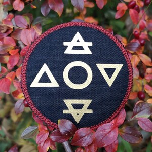 May include: A circular black patch featuring gold elemental symbols: air, water, fire, and earth. It has a red stitched border. The background shows vibrant red and orange autumn leaves.