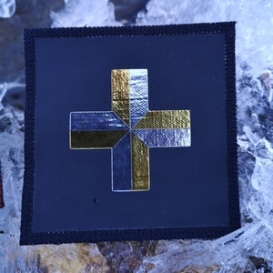May include: A black patch with a gold and blue cross design. The cross is made of fabric and has a textured look.