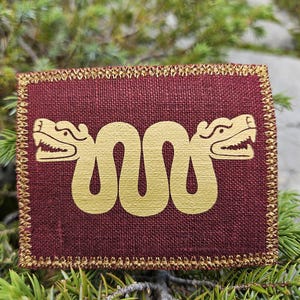May include: A square-shaped patch with a deep burgundy linen-like fabric base. The center features a metallic gold screen-printed design of a double-headed serpent with stylized features. The edges are finished with a gold-colored stitched border.