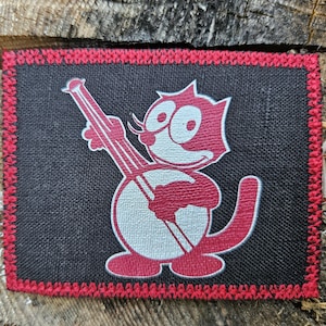 May include: A rectangular black fabric patch featuring a red and white graphic of a cartoon cat playing a banjo. The patch has a distinct red stitched border around the entire perimeter, providing a textured finish to the woven material.