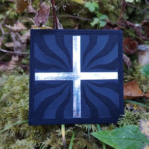 May include: A black fabric patch with a silver cross in the center. The cross is surrounded by a swirling pattern.