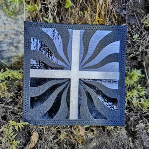 May include: A black and white fabric patch with a white cross on a black background. The background has a swirling pattern.