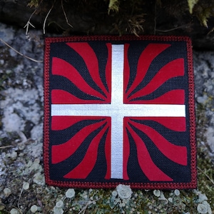 Könnte beinhalten: Ein roter und weißer gestickter Aufnäher mit einem weißen Kreuz auf schwarzem Hintergrund. Das Kreuz ist von roten Strahlen umgeben, die vom Zentrum ausgehen.