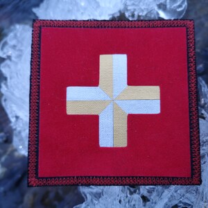 Op de afbeelding: Een rode stoffen patch met een wit kruis omrand met goud. Het kruis is gemaakt van meerdere stofdelen die aan elkaar zijn genaaid. De patch heeft een zwarte en rode rand.