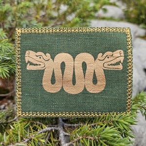 Può includere: Una toppa rettangolare in tessuto verde scuro con un disegno ricamato in filo dorato metallizzato che raffigura un serpente a due teste. La toppa è rifinita con un bordo decorativo cucito in filo dorato su tutti i lati, creando contrasto.