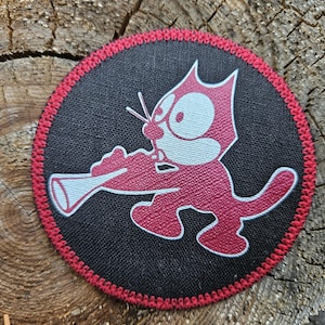 May include: A circular embroidered patch featuring a black background with a red border. The design depicts a stylized cartoon cat in red and white, playing a trumpet. The patch has a textured fabric appearance and a stitched edge finish.