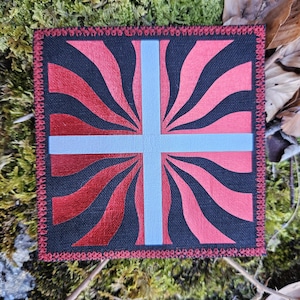 May include: Square patch with a design featuring a light blue cross over a red and black pattern. The patch has a red border and is set against a mossy background.