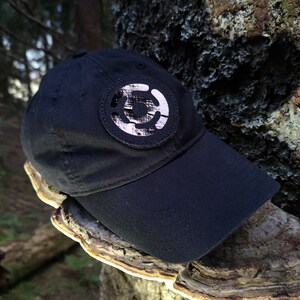 May include: A black baseball cap with a circular patch featuring a white and gray design. The cap is made of a durable fabric and has a curved brim. The cap is resting on a tree trunk.