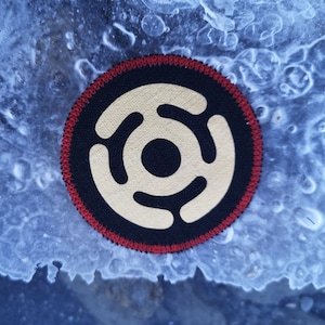 Peut inclure: Un patch rond noir et blanc avec une bordure rouge. Le patch a un design stylisé de trois cercles concentriques avec un cercle central.