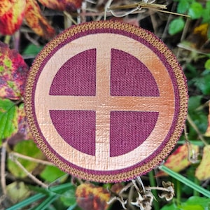Puede incluir: Un parche redondo bordado con una cruz marrón dentro de un círculo sobre un fondo burdeos. El parche tiene un borde dorado con un detalle de costura marrón.