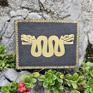 Puede incluir: Un parche rectangular de tela negra con un gráfico de una serpiente de dos cabezas de color dorado en el centro. El parche está bordeado por un grueso borde bordado en hilo dorado metálico, creando un contraste marcado con el fondo oscuro.