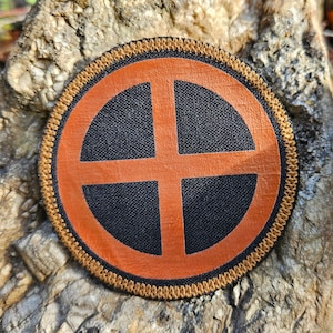 Könnte beinhalten: Ein brauner und schwarzer gestickter Aufnäher mit einem Kreisdesign. Der Kreis hat ein braunes Kreuz darin.