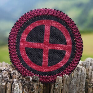 May include: A round, red and black fabric patch with a cross inside a circle. The patch is surrounded by a decorative border of red stitching.