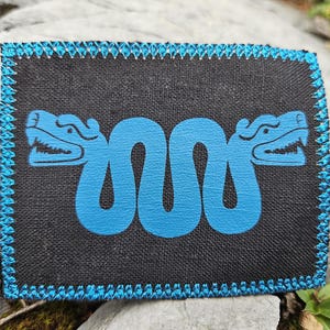 Puede incluir: Un parche rectangular de tela negra con un gráfico azul brillante de una serpiente de dos cabezas con un cuerpo ondulado. Los bordes del parche están rematados con un borde decorativo de puntada en zigzag azul brillante que enmarca el diseño.