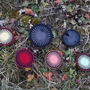May include: Six round coasters with a sunburst design in various colours, including black, white, red, yellow, and pink. The coasters are arranged on a bed of green grass and brown leaves.