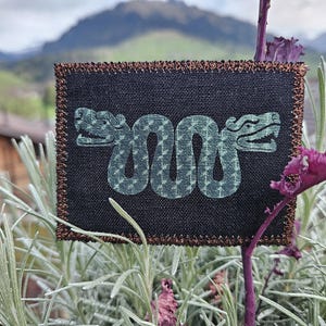 May include: A black fabric patch with a brown stitched border. The patch features a green snake design with a patterned body.