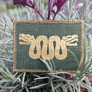 Peut inclure: Un patch en tissu vert avec un motif brodé en or de deux serpents entrelacés avec des têtes face à face.