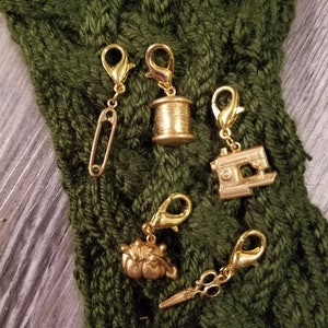 May include: Five gold-colored stitch markers with different charms. The charms include a spool of thread, a sewing machine, a pair of scissors, a safety pin, and a small crab.
