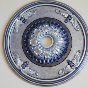 May include: A decorative ceiling medallion with a silver and blue color scheme. The circular design features ornate floral and leaf patterns, with a central opening. The outer edge and inner details are accented with a darker blue.