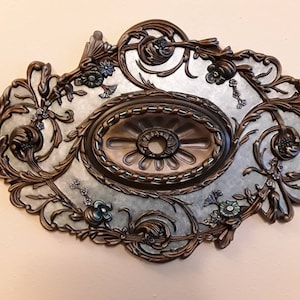 May include: A decorative ceiling medallion with an ornate, floral design. The medallion is made of metal and has a silver and bronze finish. It features a central oval with a raised, circular design.