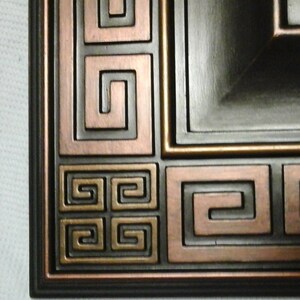 May include: A close-up of a dark brown picture frame with a copper-colored Greek key design. The frame has a raised edge and a beveled surface.
