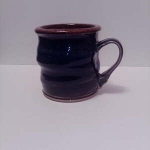 May include: A handmade ceramic mug with a dark blue exterior and a brown rim and base. The mug has a curved handle and a slightly textured surface. The mug is approximately 4 inches tall.