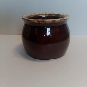 May include: A small, round, dark brown ceramic pot with a lighter brown rim. The pot has a glossy finish and a simple, classic design. It could be used for storage or as a decorative item.