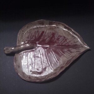 Puede incluir: Un plato de cerámica con forma de hoja y acabado brillante. El plato presenta un centro de color marrón rojizo intenso que se transforma en tonos marrones más claros y apagados hacia los bordes. Incluye un tallo esculpido y venas visibles.