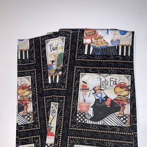May include: Set of three black and white kitchen towels with a gold and white border. The towels feature a chef character and the words "Let's Eat", "Bonjour", and "Fiesta".