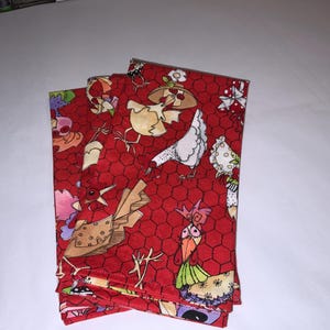 May include: A stack of four red fabric napkins with a white chicken pattern. The chickens are wearing hats and have different expressions. The background is a red honeycomb pattern.