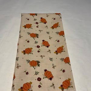 May include: Folded beige fabric with a repeating pattern of orange pumpkins, green leaves, and small burgundy flowers. The fabric has a light, natural color and is likely a decorative textile for autumn.