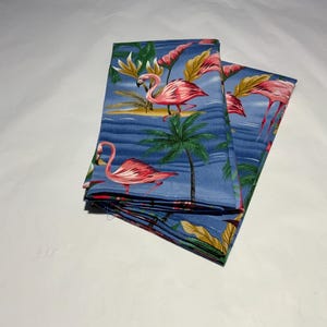 May include: Blue fabric napkins with a tropical flamingo design. The napkins feature pink flamingos, palm trees, and other tropical foliage against a blue background. The napkins are folded and stacked.