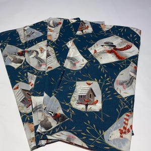 May include: Four folded cloth napkins with a winter-themed pattern. The dark teal fabric features a repeating design of snowmen, birds, and birdhouses. The napkins are made of a soft, likely cotton, material.