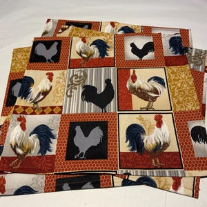 May include: A stack of four square fabric placemats. Each placemat features a repeating pattern of rooster illustrations in various colors and styles. The roosters are set against a background of red, gold, and cream-colored squares.