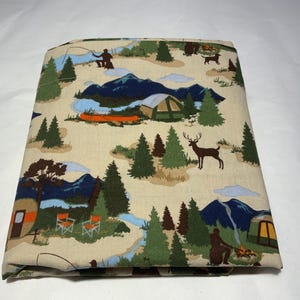 May include: A folded fabric with a camping theme. The design features a beige background with illustrations of mountains, trees, tents, a canoe, a deer, and people fishing and camping. The color palette includes green, blue, brown, and orange.