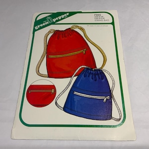 May include: A sewing pattern envelope for a string bag by The Green Pepper, labeled F865. The cover features illustrations of a red drawstring backpack and a blue drawstring backpack, both with front zipper pockets and tan cord straps.