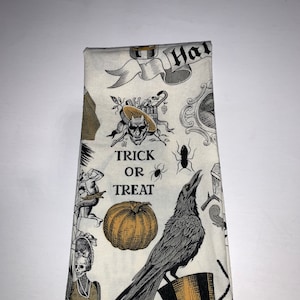 May include: A white kitchen towel with a black and gold Halloween print. The print features a crow, a pumpkin, a spider, and the text "Trick or Treat".