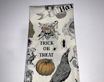 May include: A white kitchen towel with a black and gold Halloween print. The print features a crow, a pumpkin, a spider, and the text "Trick or Treat".