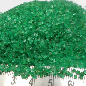 May include: A close-up of a pile of small, vibrant green emerald crystals. The crystals are irregularly shaped and appear to be raw or unpolished. A metal ruler with black markings is visible at the bottom, providing scale.