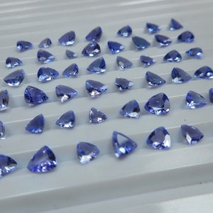 Op de afbeelding: Een verzameling gefacetteerde, driehoekige blauwe edelstenen, gerangschikt op een lichtgrijze ondergrond. De edelstenen variëren in grootte en vorm, met verschillende blauwtinten en interne helderheid. De opstelling is netjes.
