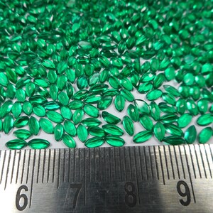 Pode incluir: Uma imagem em close-up mostra inúmeras pequenas pedras preciosas verdes esmeralda, com corte marquesa. As gemas estão espalhadas por uma superfície, com uma régua para escala. As pedras facetadas refletem a luz. Adequado para fazer joias.