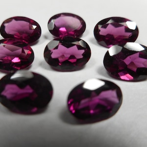 May include: Eight oval-cut, faceted gemstones in varying shades of deep magenta and purple. The gemstones are arranged on a white surface, with light reflecting off their cut surfaces. These gemstones are suitable for jewelry making.