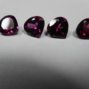May include: Four heart-shaped gemstones in a deep magenta hue. The faceted surfaces reflect light, creating highlights. The gems are arranged on a white surface, showcasing their polished appearance. Suitable for jewellery.