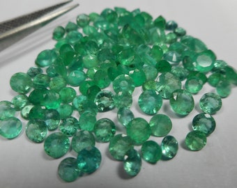 May include: A selection of faceted emerald gemstones in diverse shapes and sizes, including round, oval, and pear cuts. The gemstones are a vivid green hue and are spread across a white surface. Silver tweezers are visible in the upper left corner.